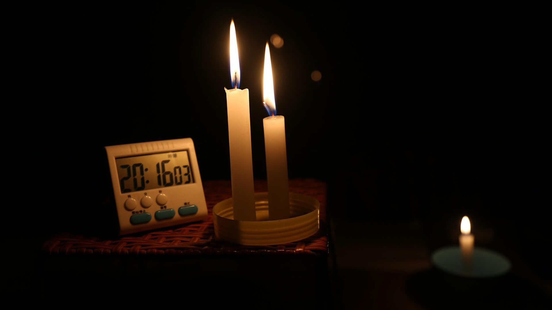 Candles burning near a digital clock in a dark room.