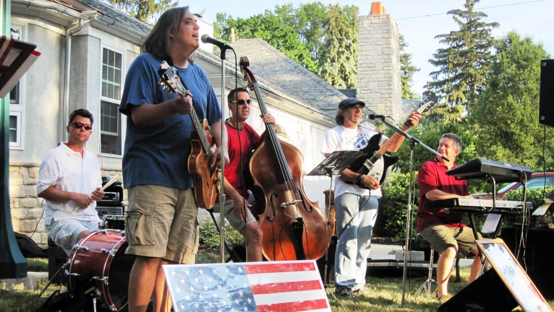 Band performing outdoors, featuring guitar, bass, drums, keyboard. US flag visible.