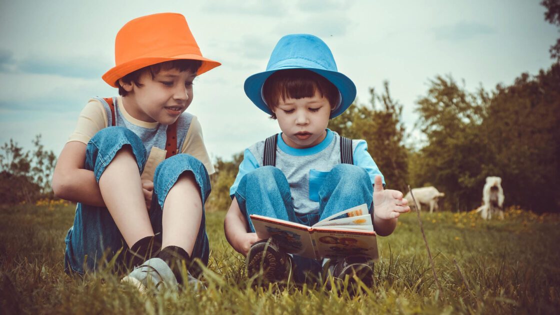 Two kids in hats read a book together in a grassy field with goats in the distance.