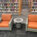 Library seating area with orange chairs and a gray table with dual outlets. Bookshelves in the background.