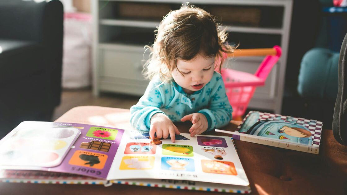 A toddler in blue pajamas points at colorful shapes in a children's book.