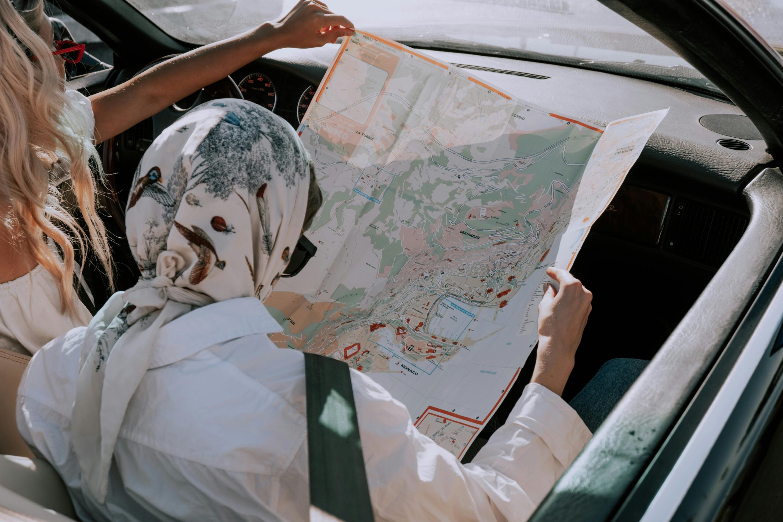 Two women in a convertible sports car with lowered roof reading a map of a coastal town. Sunlight casting blue shadows.