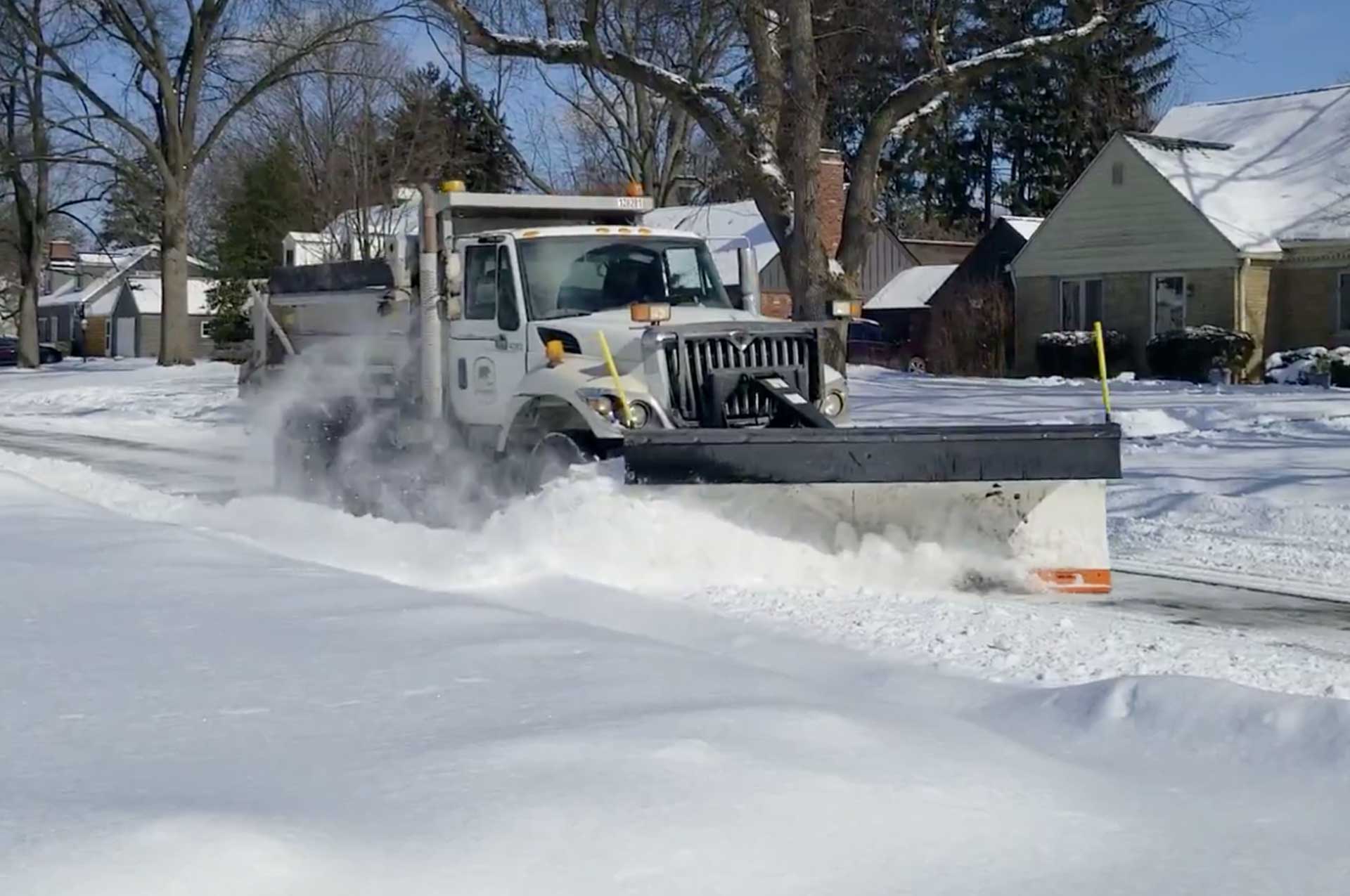 Snow plow clearing snow-covered street in a residential neighborhood.