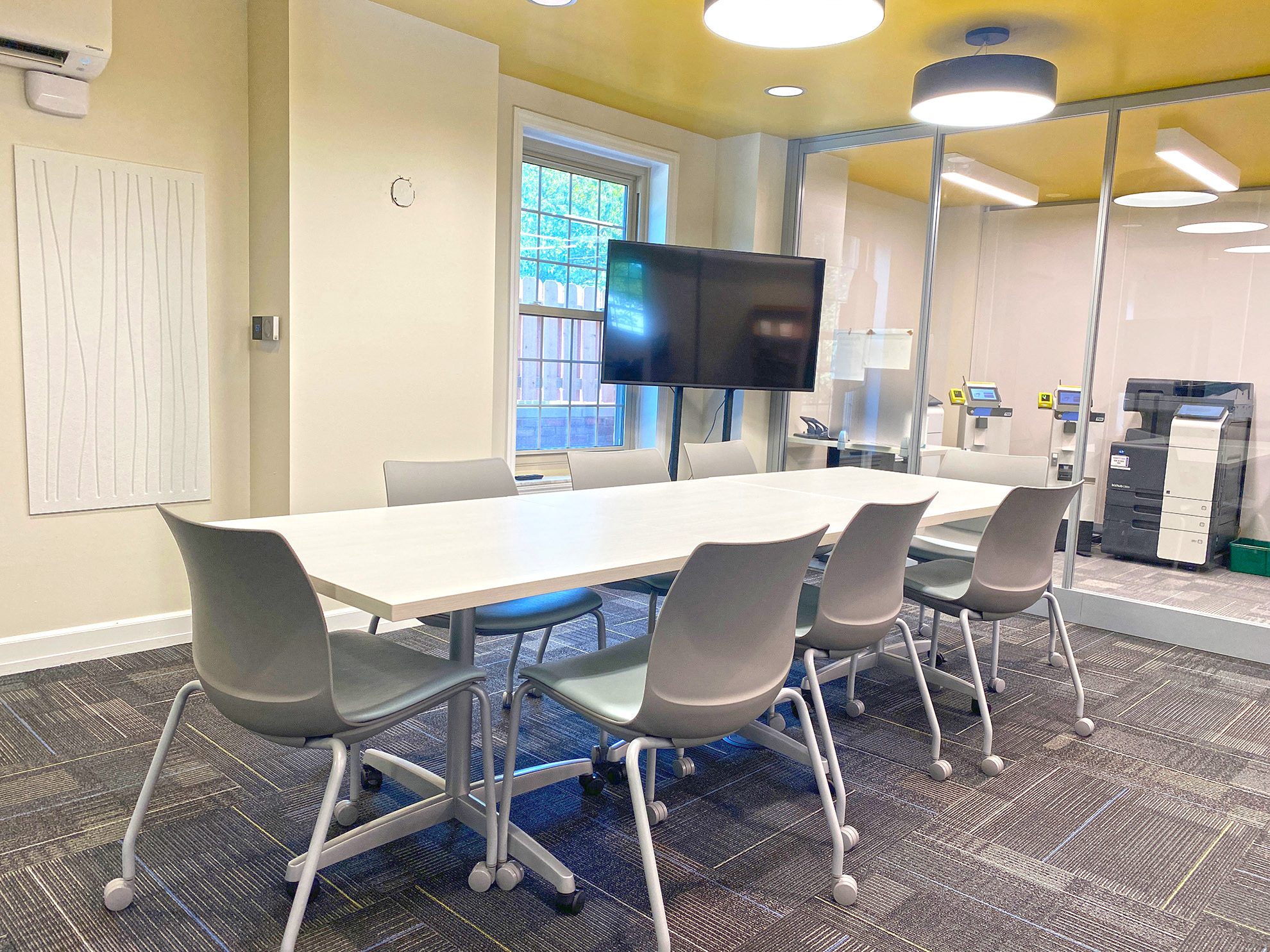 Bright meeting room with a white table, gray chairs, and a large TV on a stand.