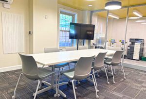 Conference room with a large table, chairs, TV on a stand, yellow ceiling and glass wall.