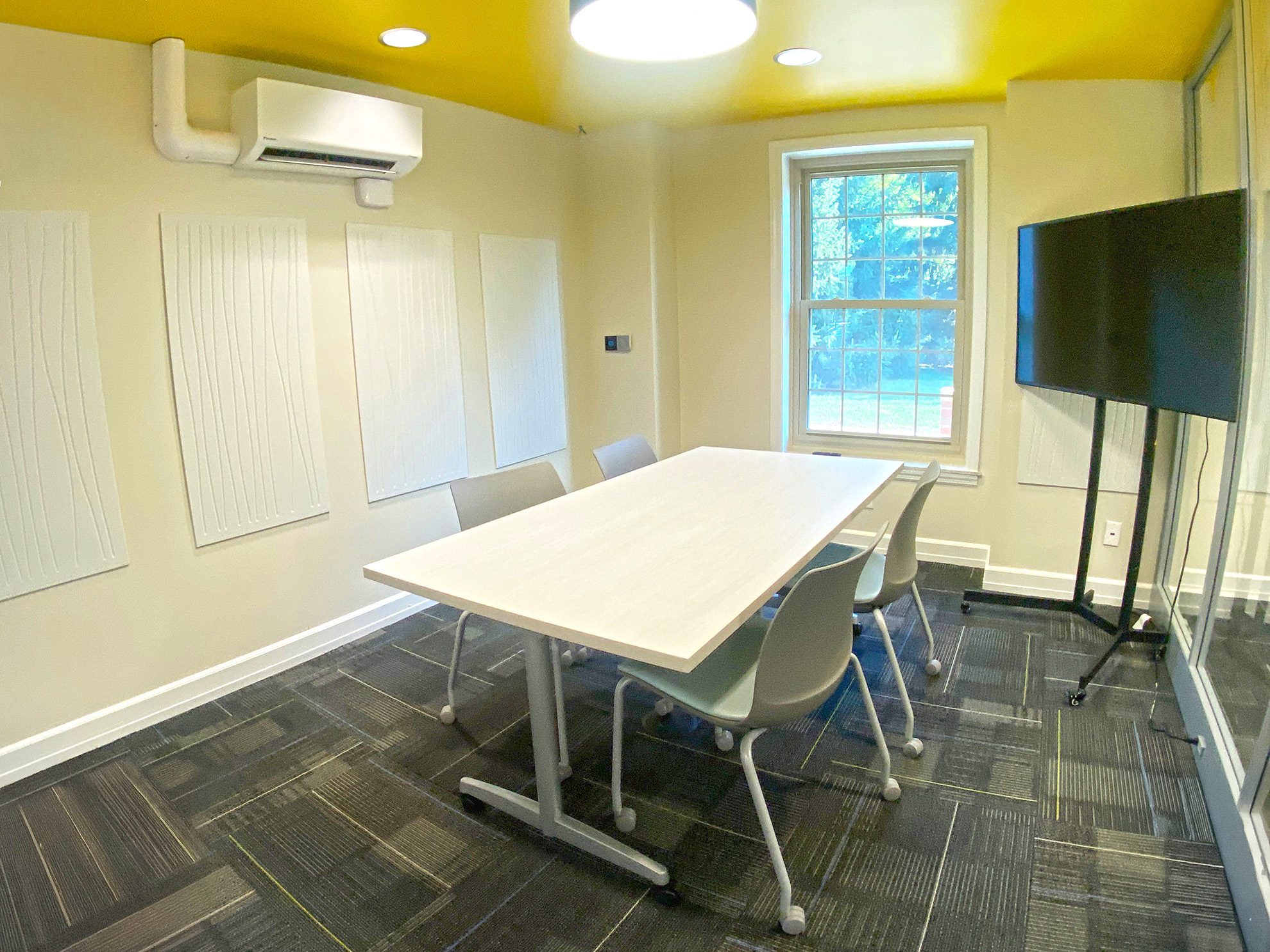 Conference room with a table, chairs, TV on a stand, yellow ceiling and glass wall.