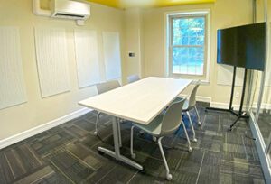 Conference room with a table, chairs, TV on a stand, yellow ceiling and glass wall.