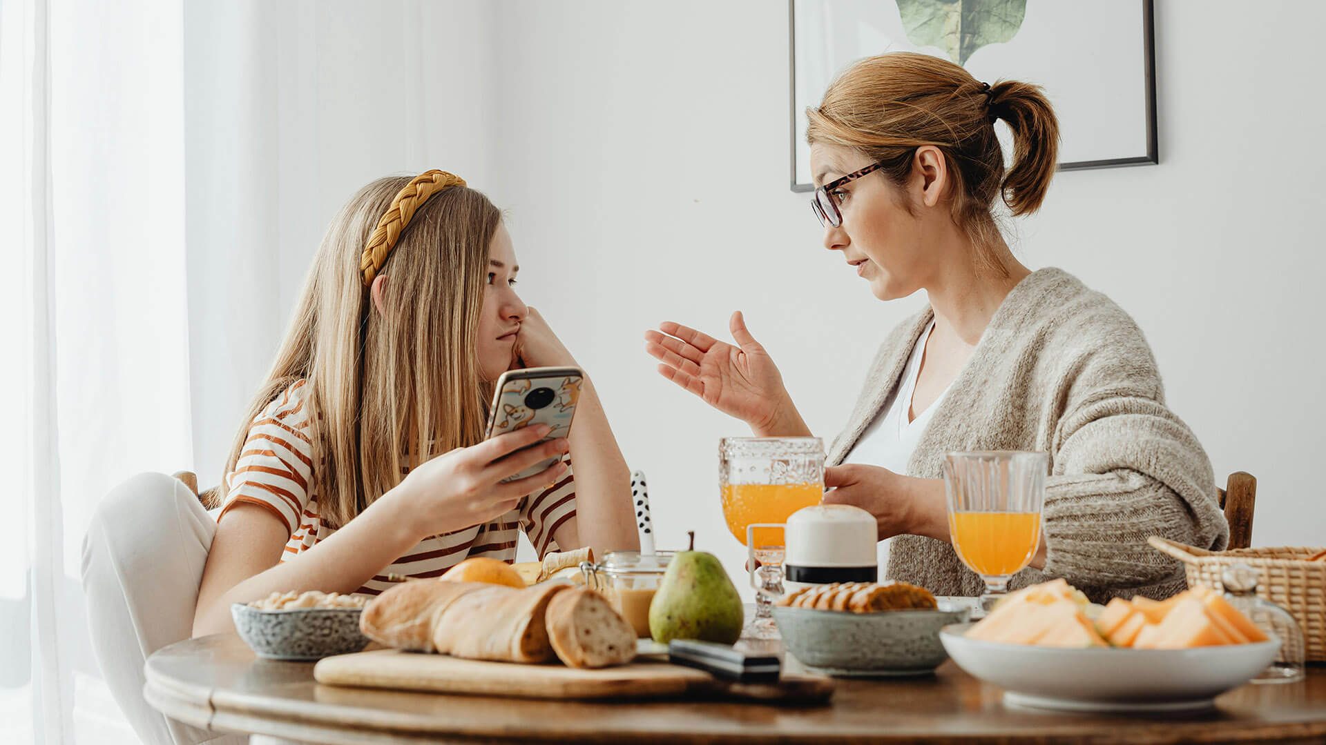 Mother talking to teen daughter distracted by phone at breakfast.
