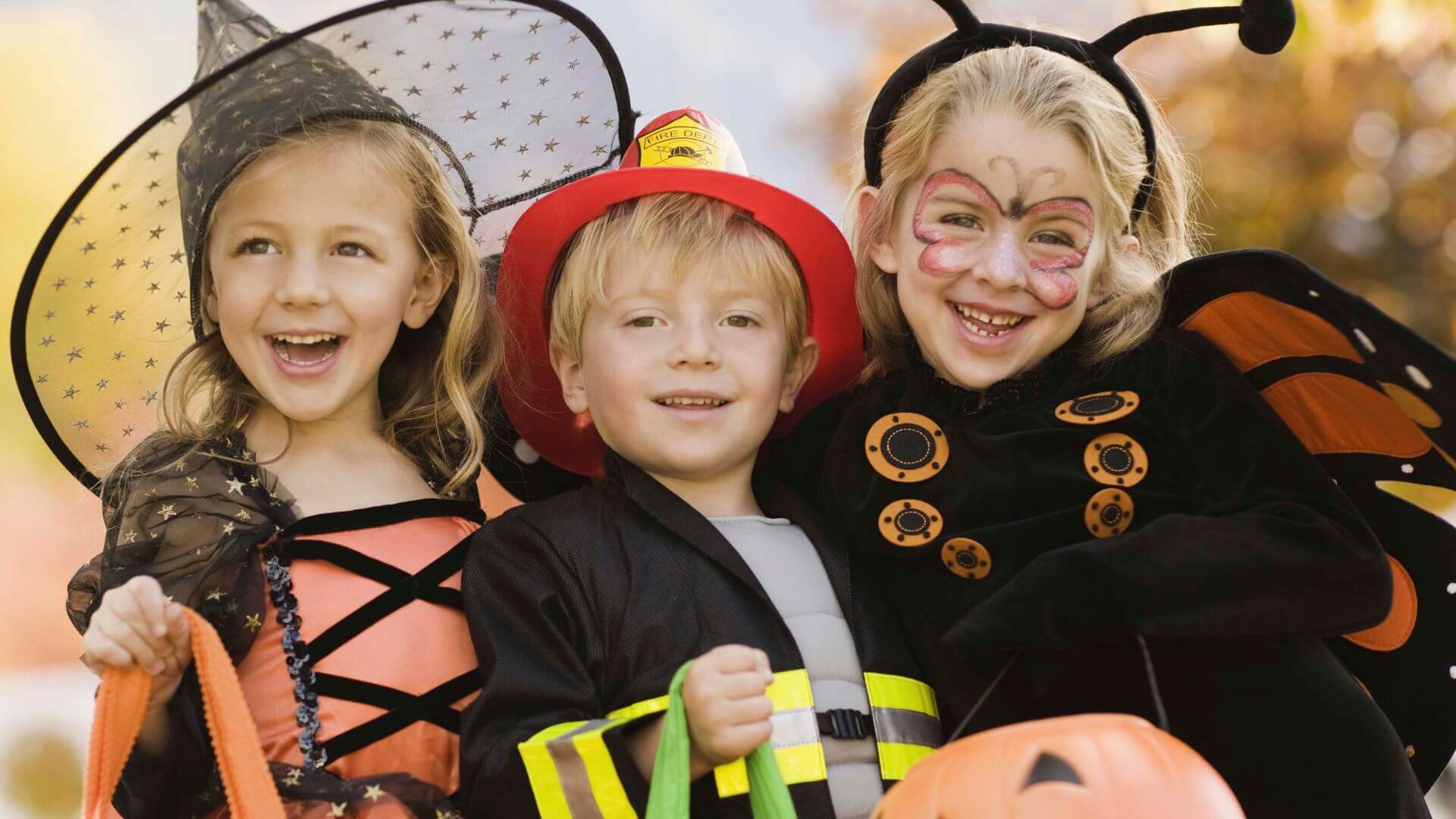 Three smiling children in Halloween costumes: witch, firefighter, and butterfly.