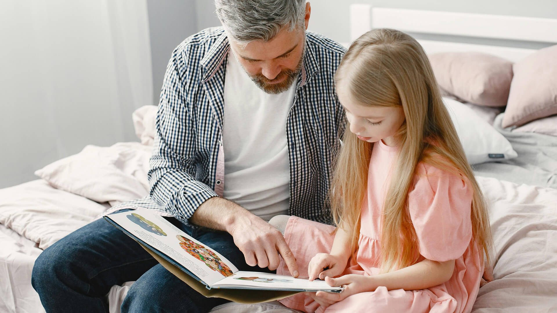 A dad reading with his daughter sitting on a bed.