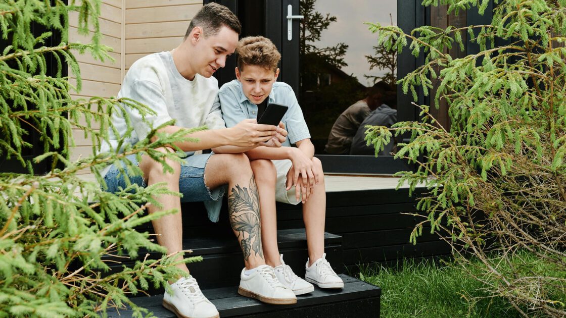 Two people, an adult and a teenager, sit on steps, looking at a smartphone. One has a leg tattoo.