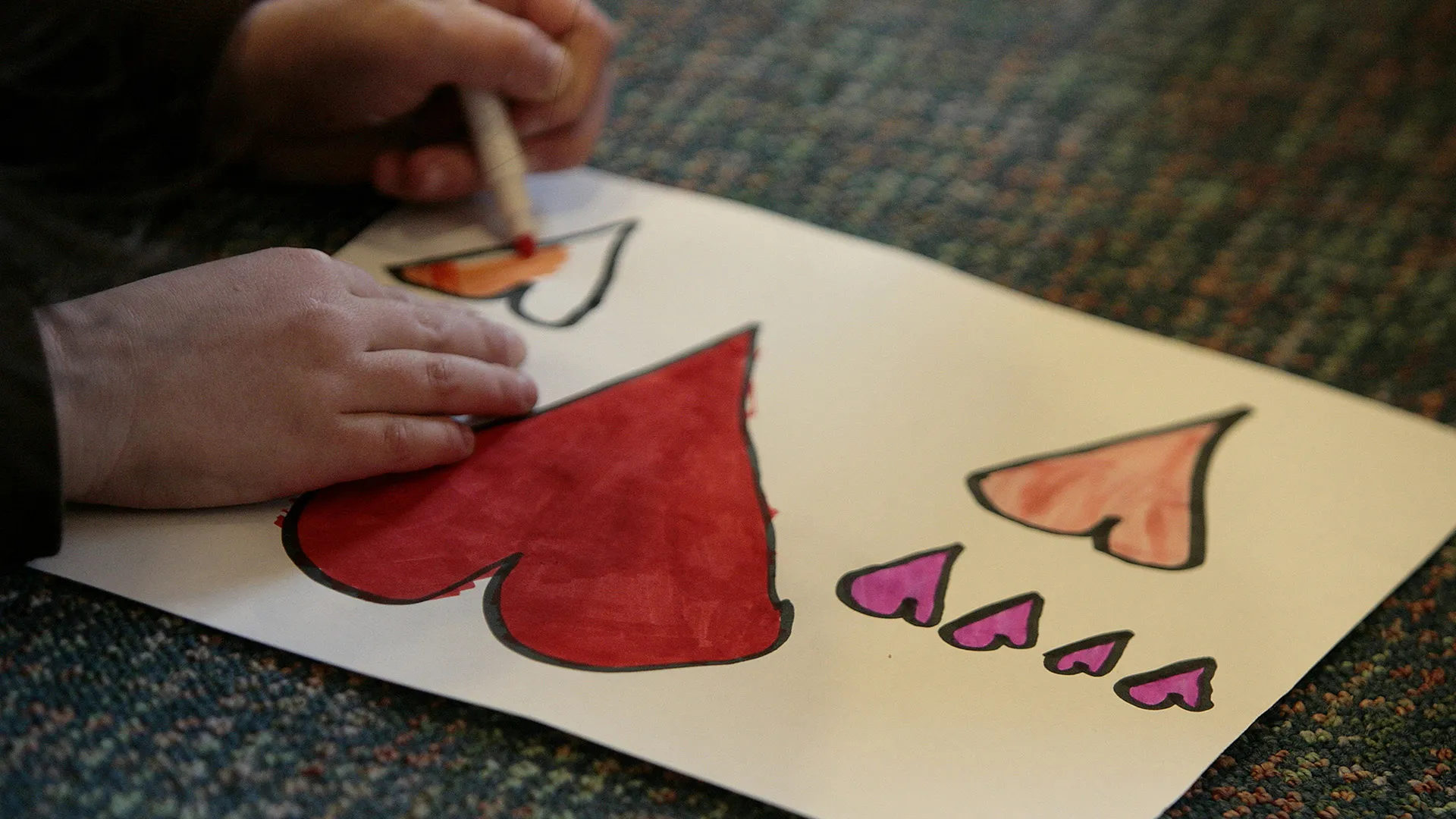 A child colors an outlined picture of colorful hearts.