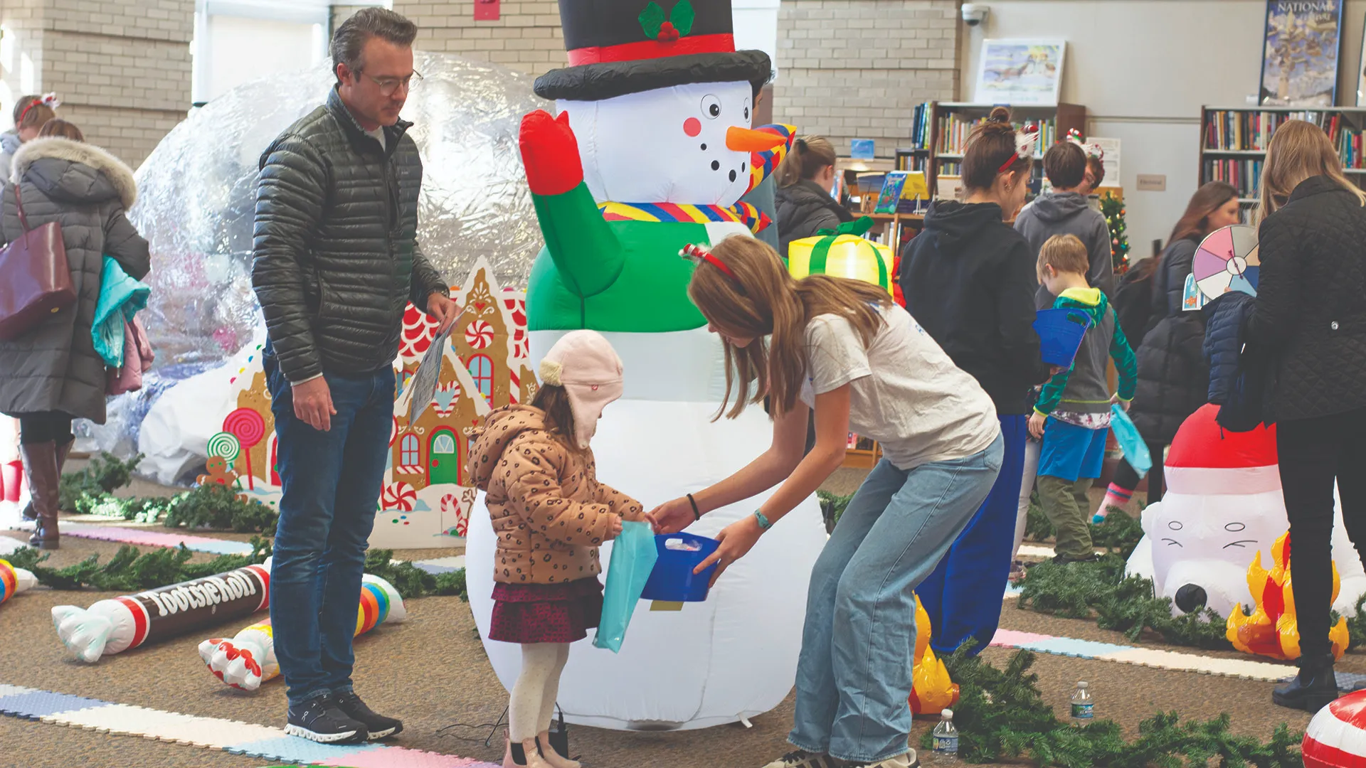 At a holiday event, a child receives a gift from an older child next to an inflatable snowman.