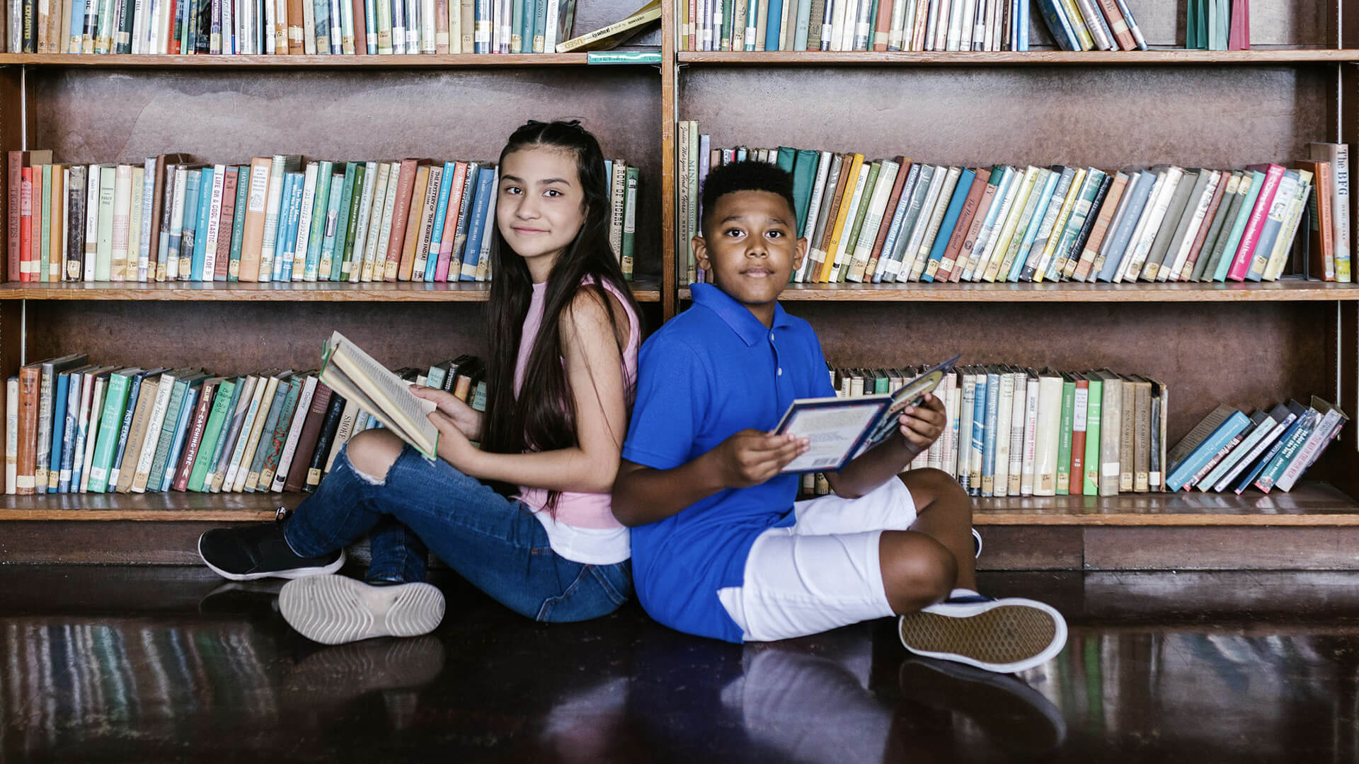 A boy and a girl reading books back-to-back against a bookshelf in a library.