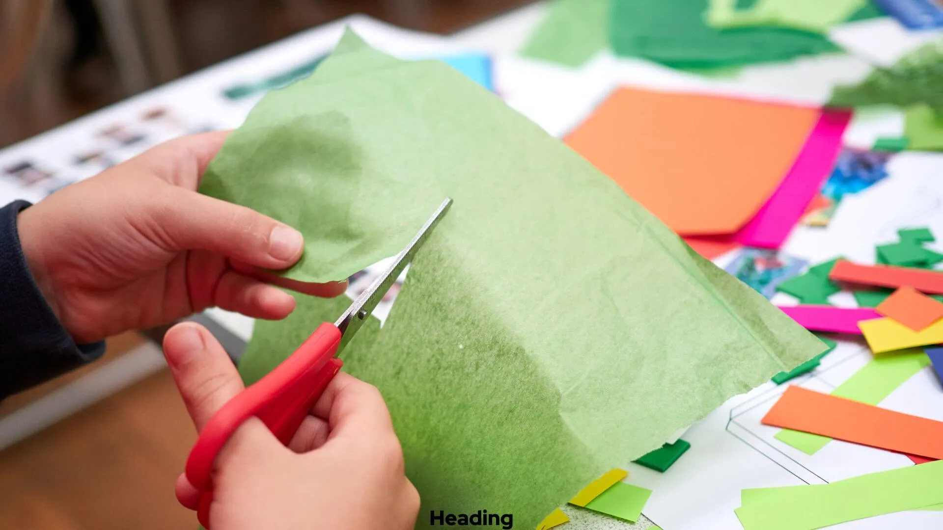 Child's hands using red scissors to cut green tissue paper. Colorful paper scraps are on the table.
