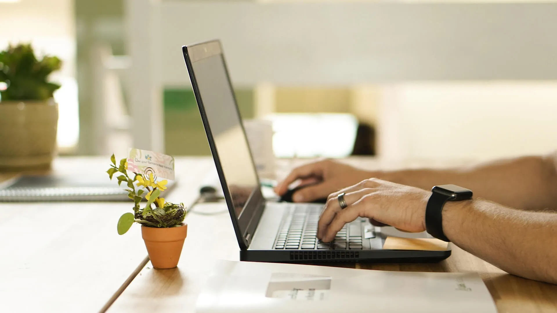 Close-up of hands typing on a black laptop on a wooden desk, with a smartwatch and a small plant.
