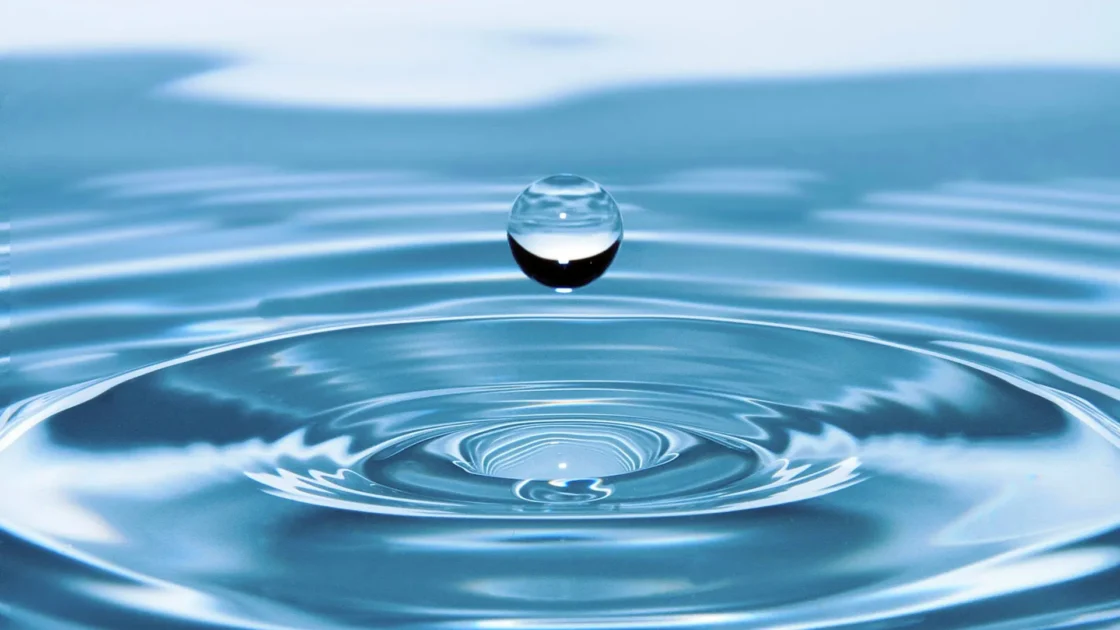 Clear spherical water drop hovering above a light blue water surface with concentric ripples.
