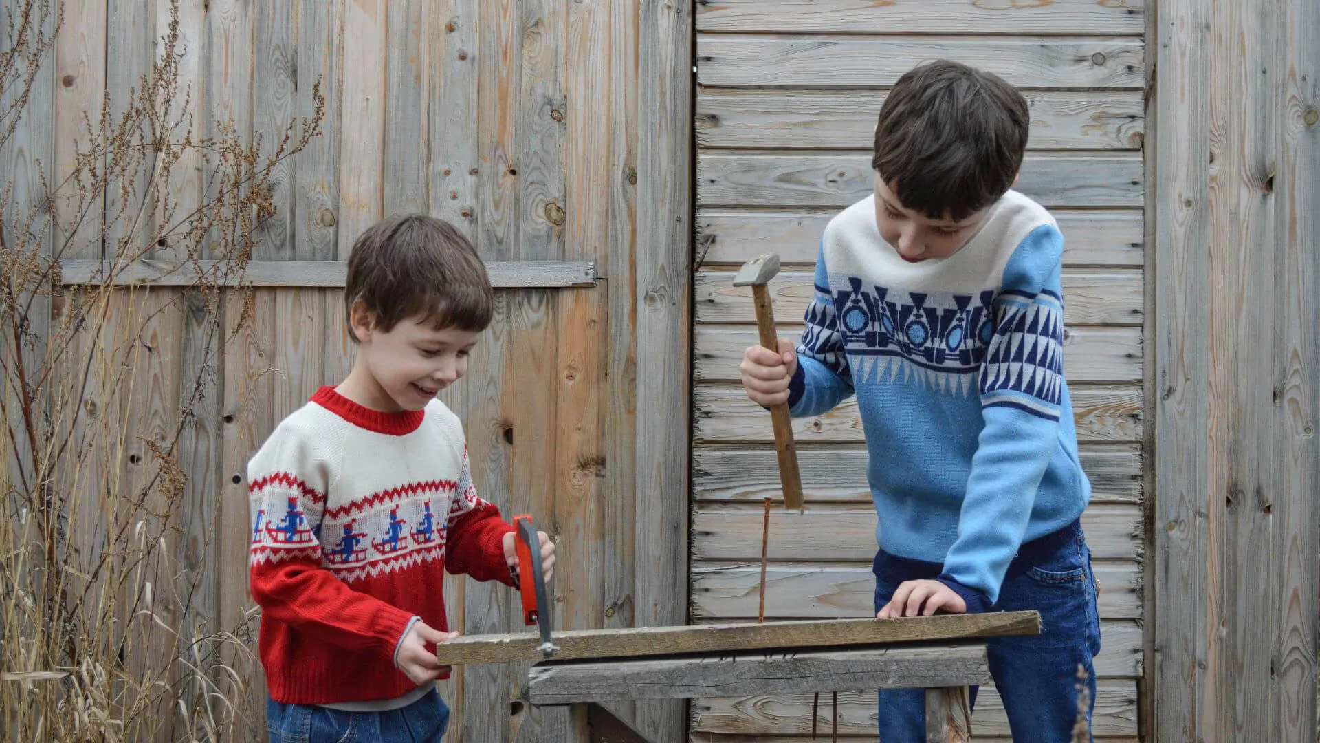 Two smiling boys in patterned sweaters sawing and hammering wood planks against a wooden fence.