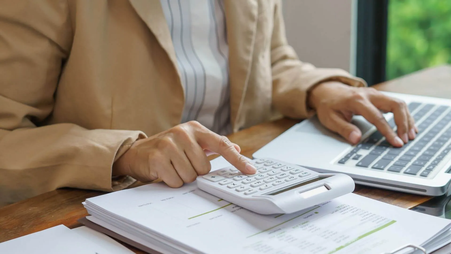 Person's hands using a calculator and laptop, with financial reports on a wooden desk.