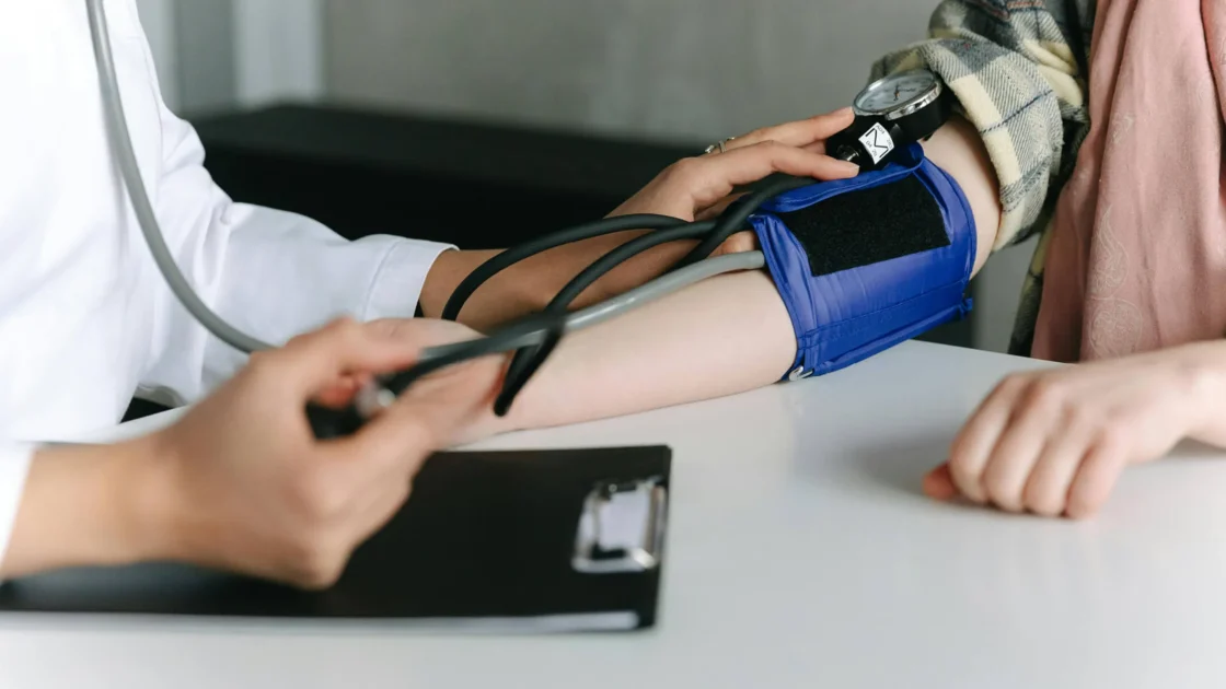 A doctor in a white coat measures a patient's blood pressure with a blue arm cuff and gauge.