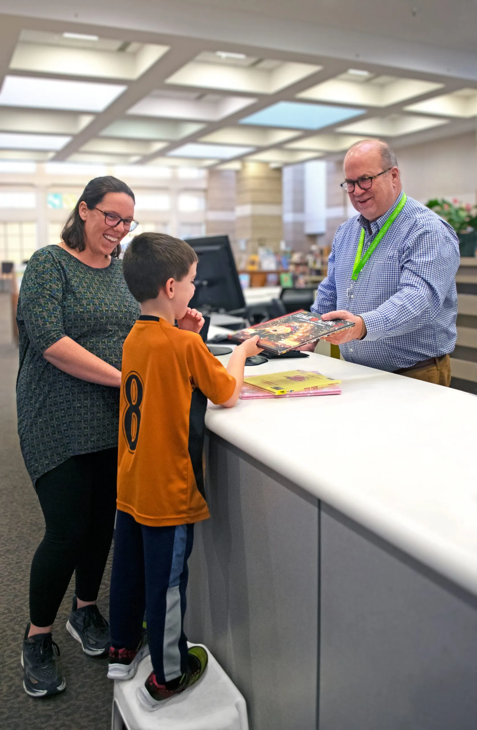 Smiling librarian hands books to a boy on a stool, as a woman watches happily in a bright library.
