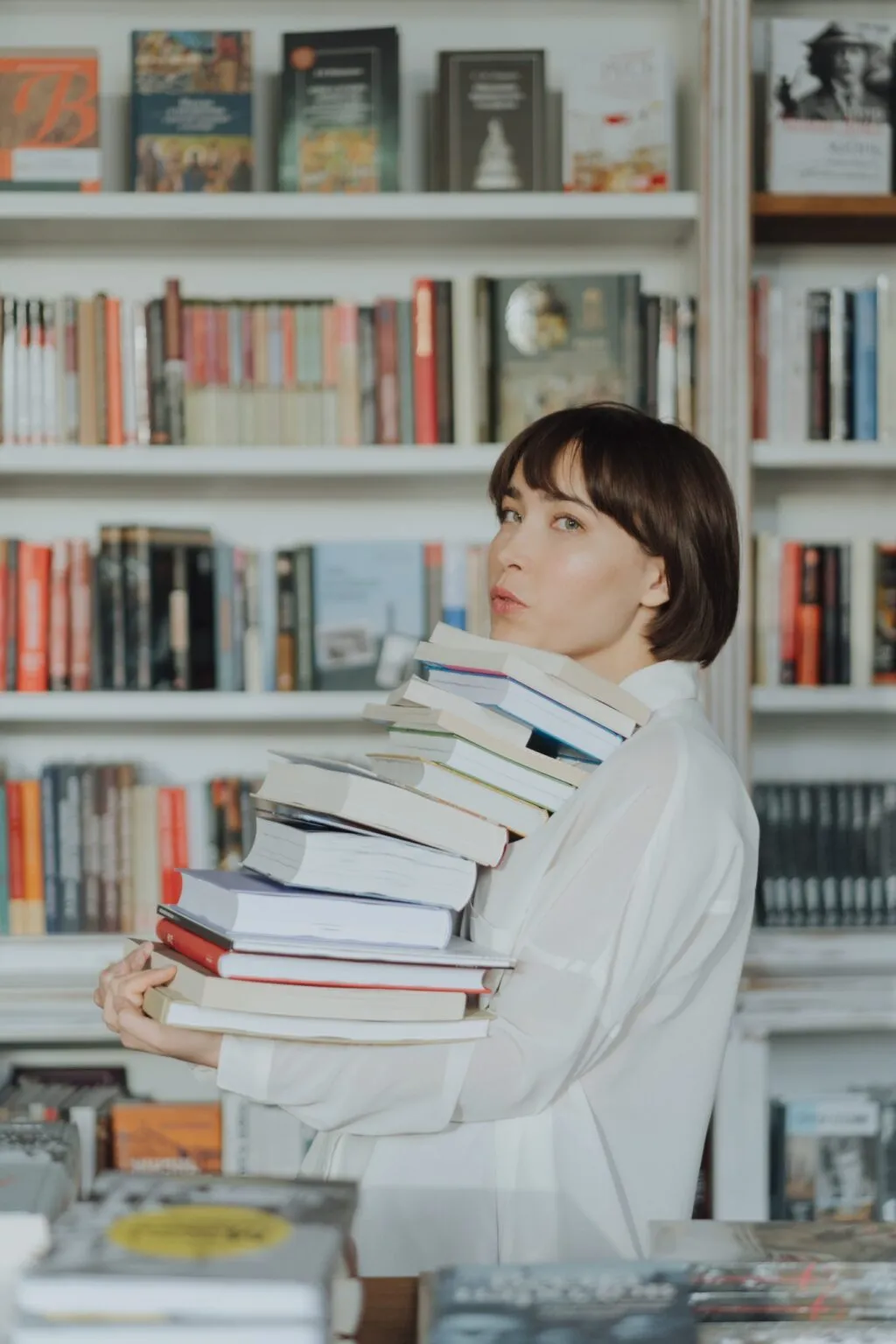 Woman in a library holds a large stack of books, looking back over her shoulder.