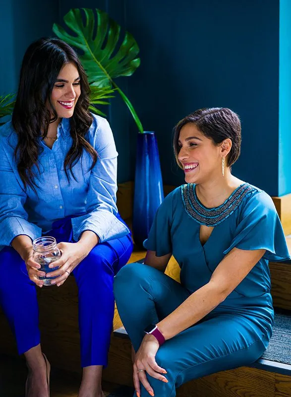 Two smiling women: one in royal blue, one in teal, chatting casually indoors.