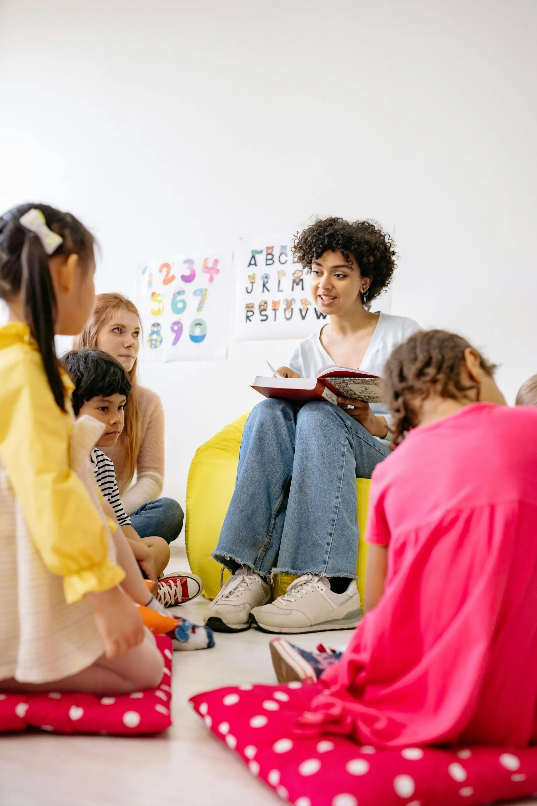 A teacher with curly hair reads a book to a diverse group of children sitting on the floor.
