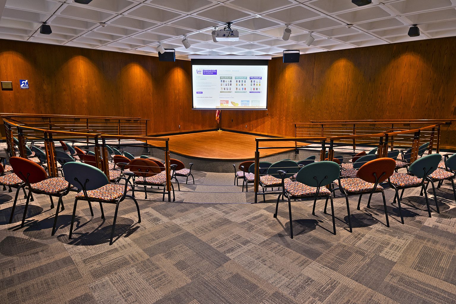 Empty tiered auditorium with colorful chairs, wood walls, and a projector displaying "Library Reads".