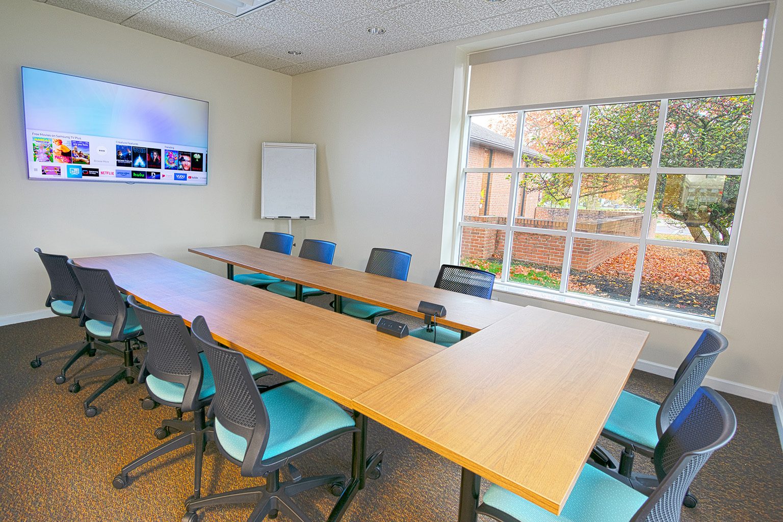 A modern conference room with a U-shaped table, teal chairs, large TV, and a window overlooking autumn trees.