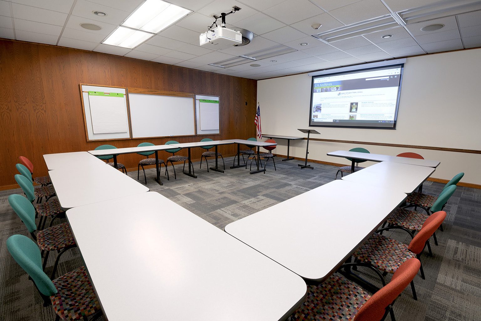 Spacious meeting room with U-shaped tables, colorful chairs, projector screen, and whiteboards.