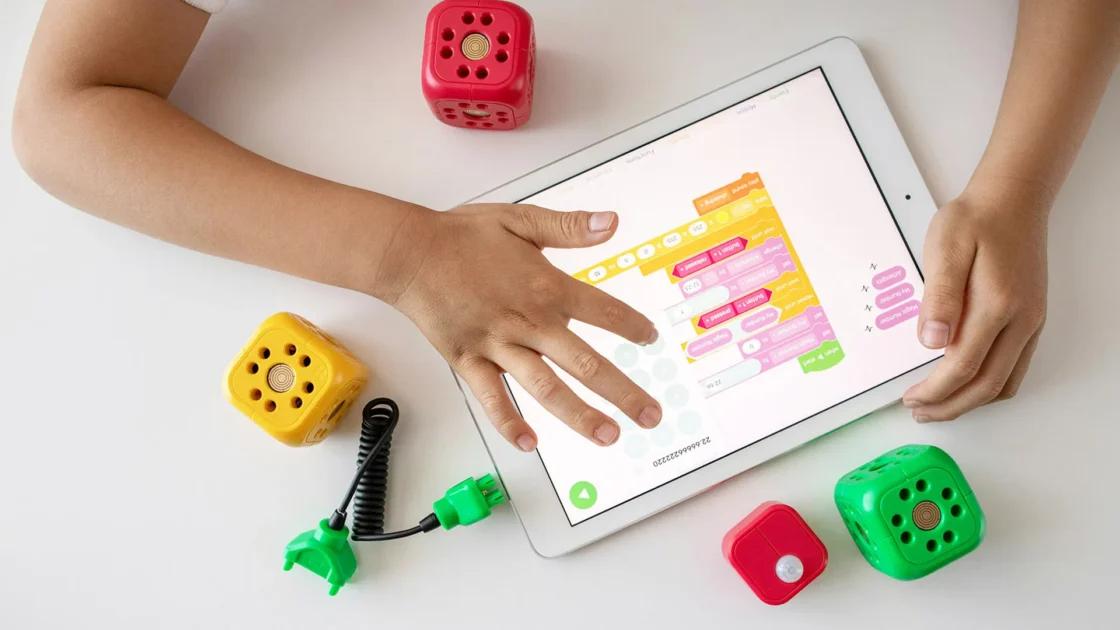 Child's hand interacts with a tablet showing block coding, surrounded by colorful robot cubes and sensors.