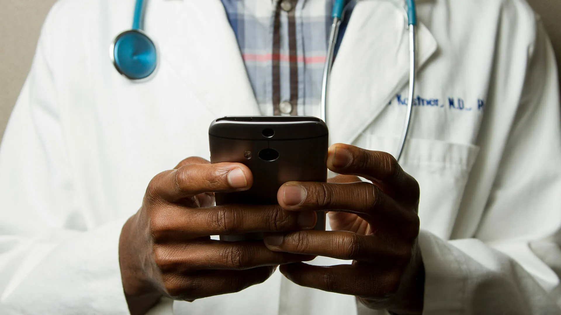 Close up of hands; doctor in a lab coat with a stethoscope, using a smartphone.