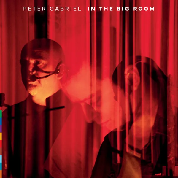 Peter Gabriel in intense red light, wearing a headset, with a blurred figure and vertical bars.