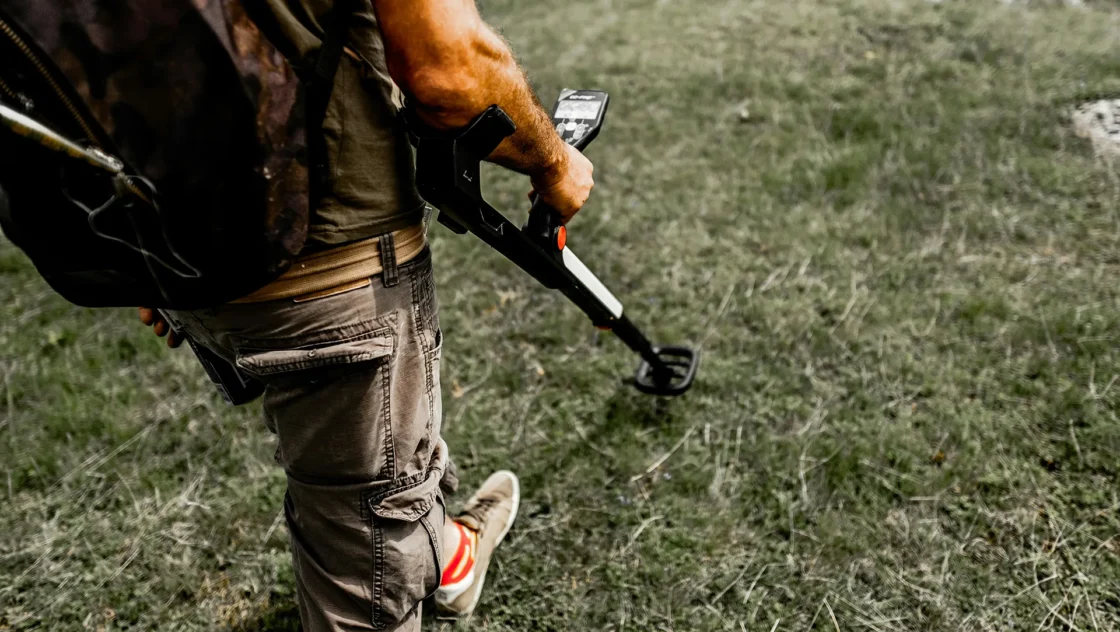 Person in cargo pants and t-shirt metal detecting in a grassy field.