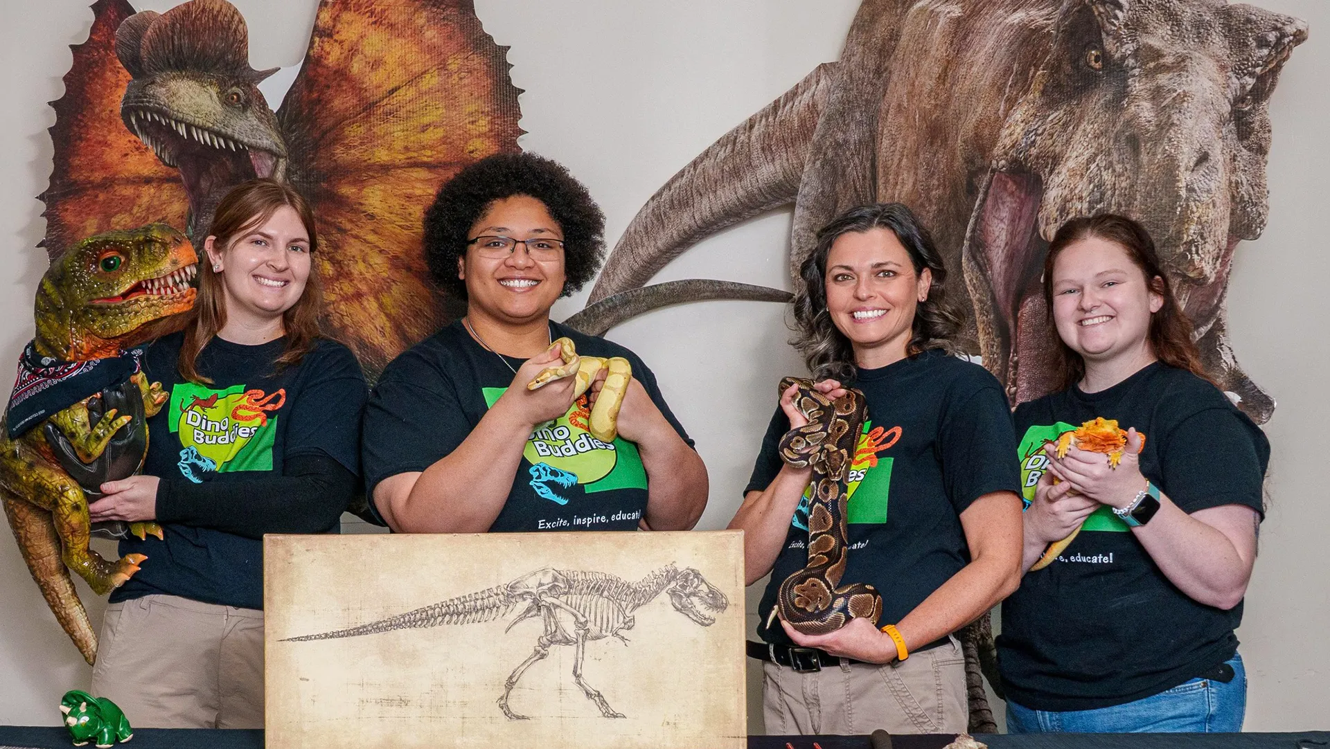 Four smiling women in Dino Buddies shirts hold snakes, a lizard, and a dino puppet, with dinosaur cutouts.