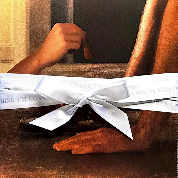 Album cover. A light-skinned hand reaches up with food toward a person on all fours, shown as two hairy arms and hands on the right. A wide white ribbon with a central bow binds them, featuring the repeating text 'I THINK I'M GONNA DIE IN THIS HOUSE'.