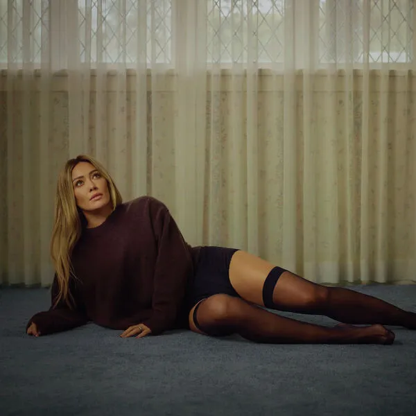 Album cover. Hilary Duff, a blonde woman, in plum sweater & black thigh-highs lies on blue carpet, looking up at curtains.