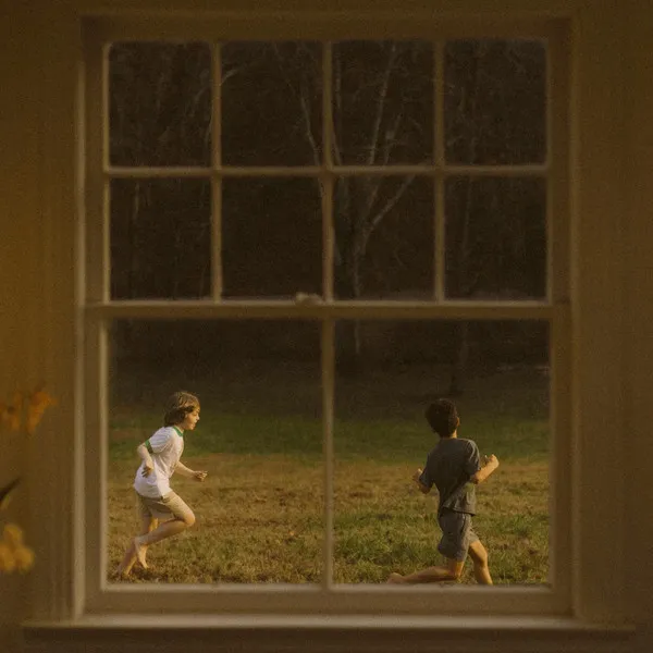Through a window, two young boys run barefoot in a field, one stumbles. Dark, bare trees behind.