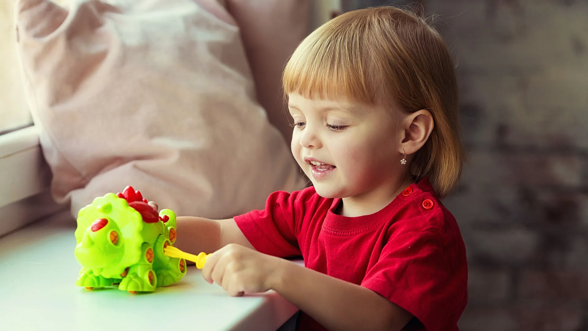 Smiling blonde girl in red plays with a green take-apart dinosaur toy using a yellow tool.