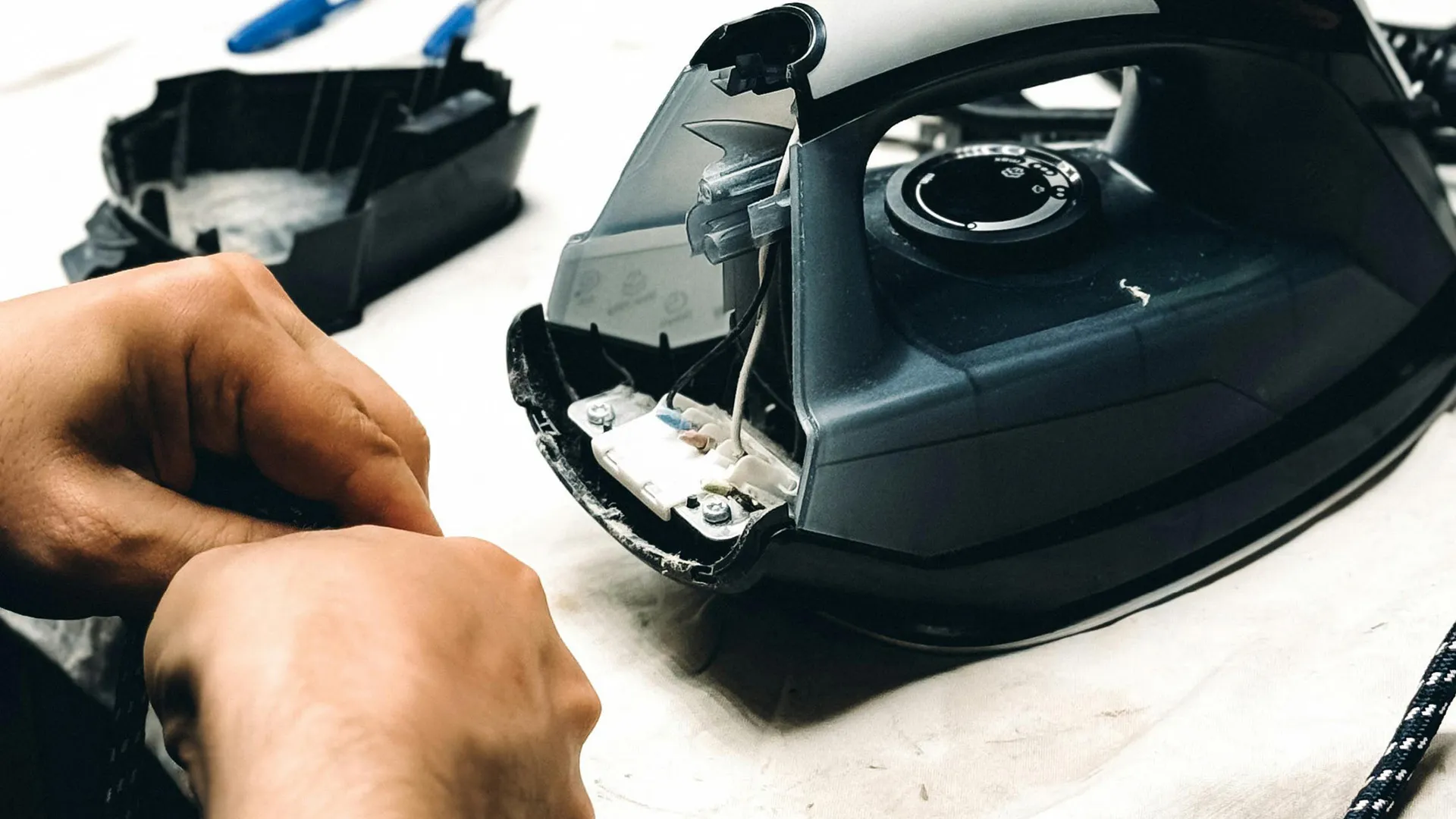 Hands repair a disassembled black clothing iron, revealing internal wires and components.