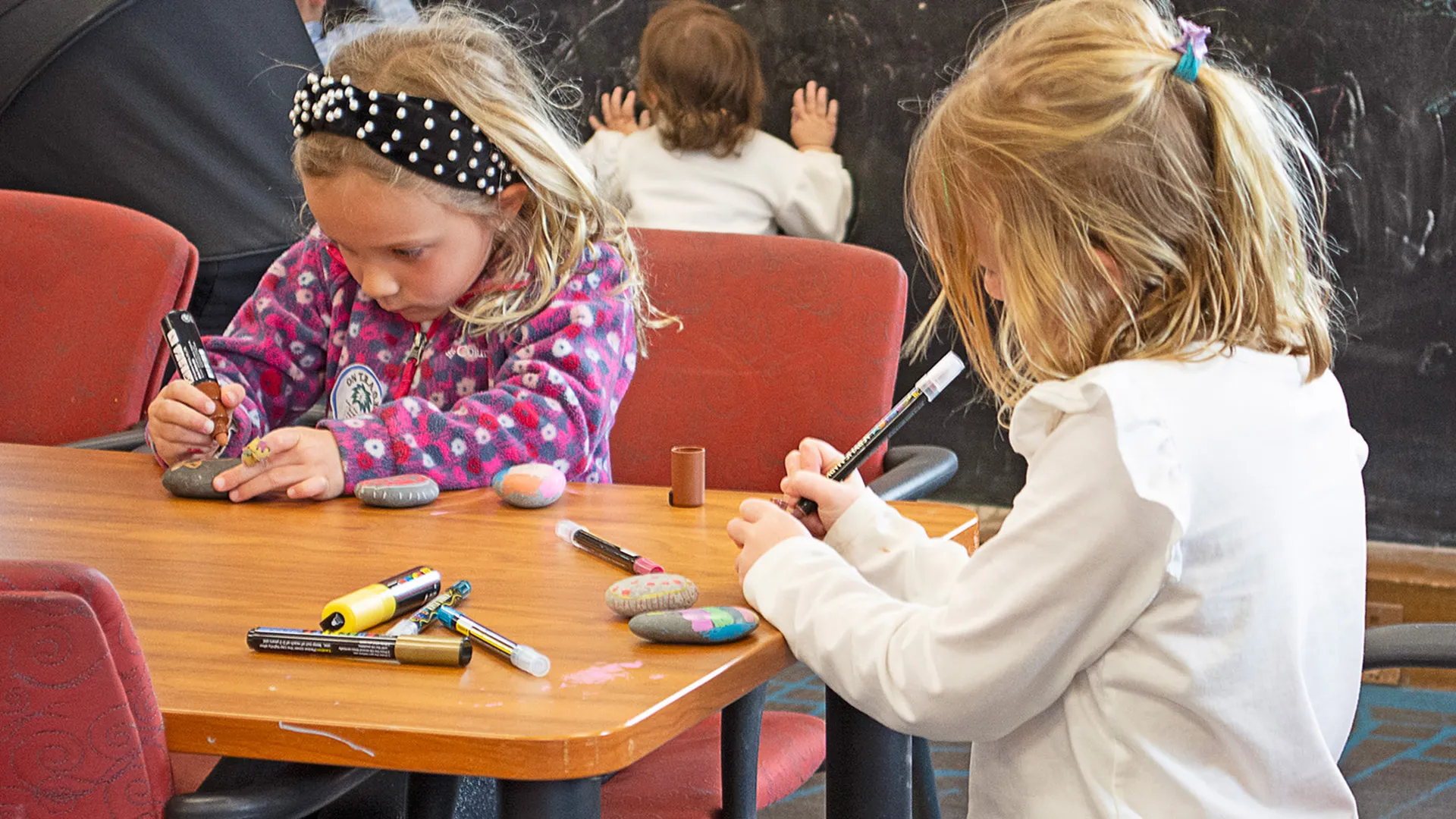 Two young girls intently decorate rocks with markers at a table. Another child is in background.