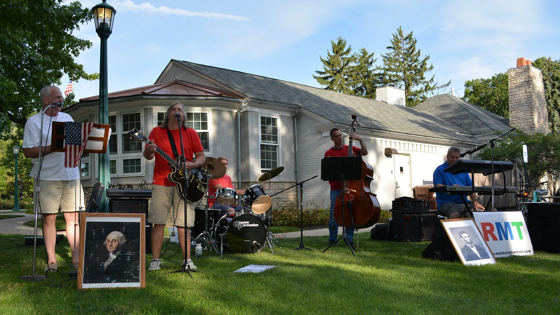Outdoor band plays on a sunny lawn with an American flag, and portraits of Washington & Lincoln.