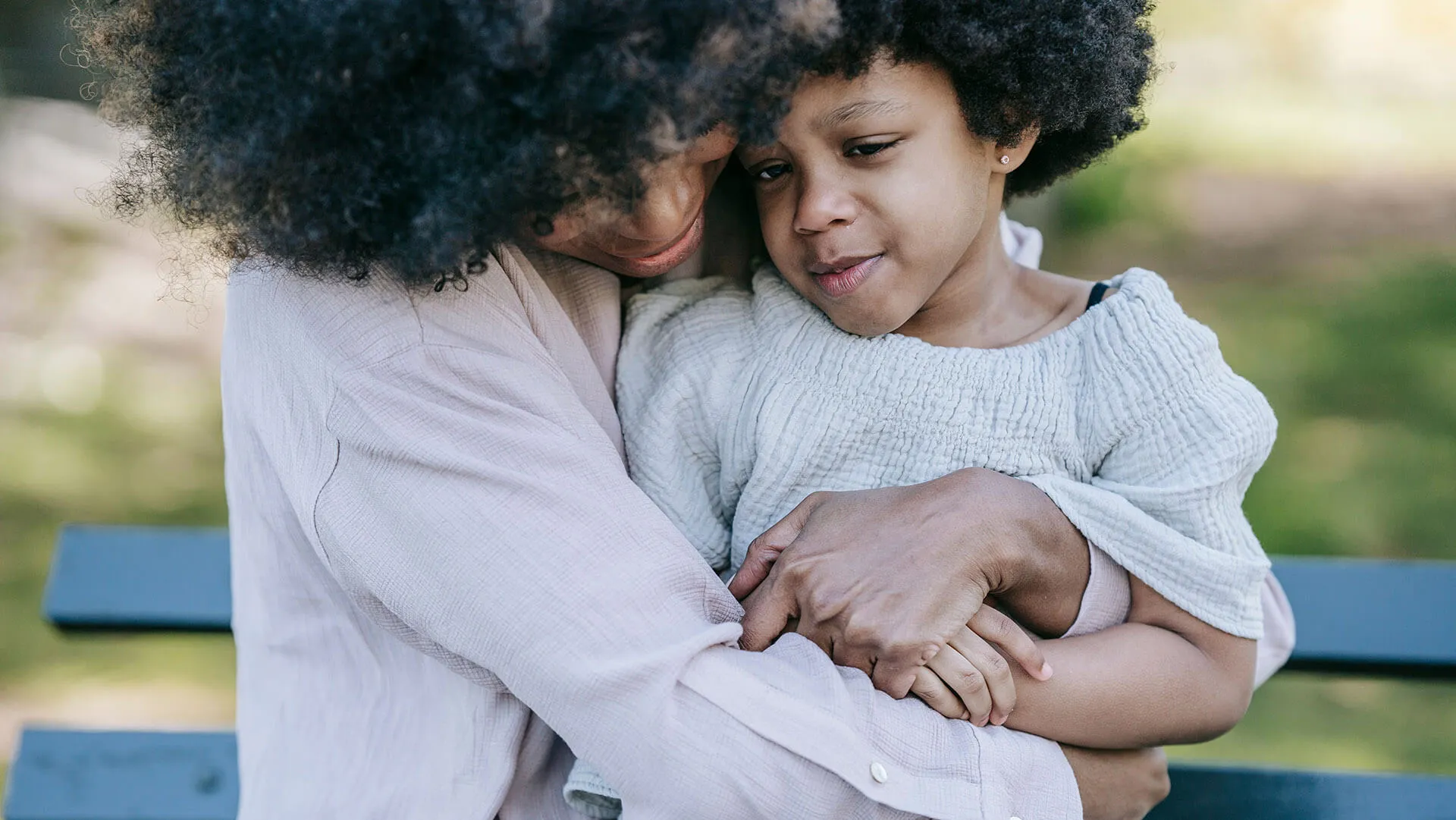 A mother with curly hair tenderly hugs her young child outdoors.