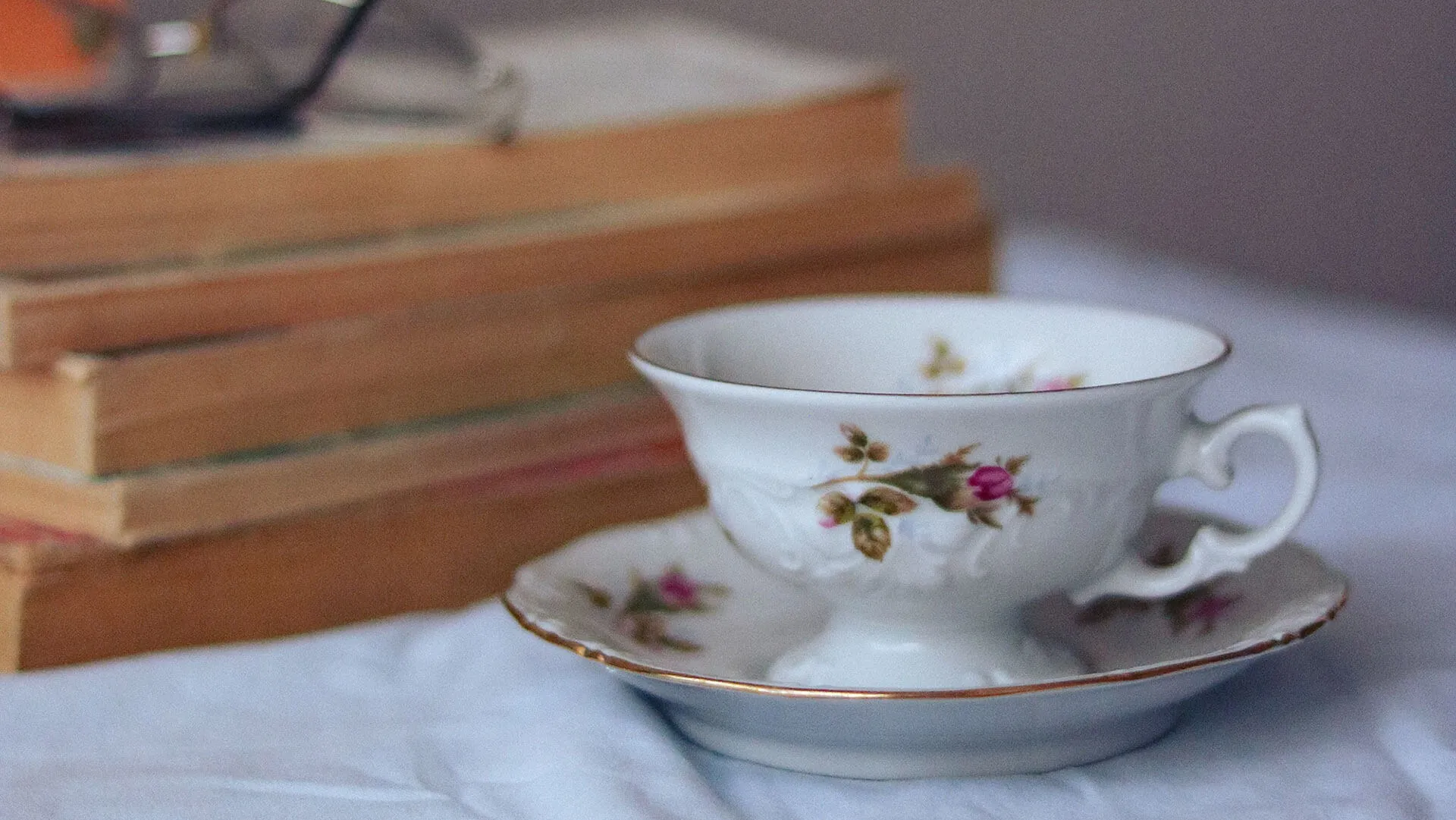A dainty floral teacup on a saucer rests near a stack of old books on a white tablecloth.