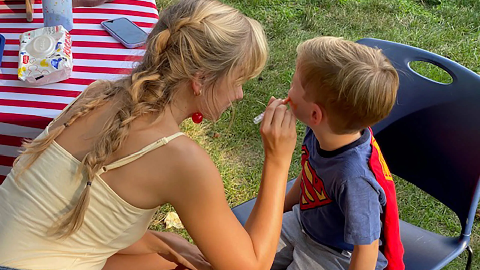 A woman with braided blonde hair face paints a young boy dressed as Superman.
