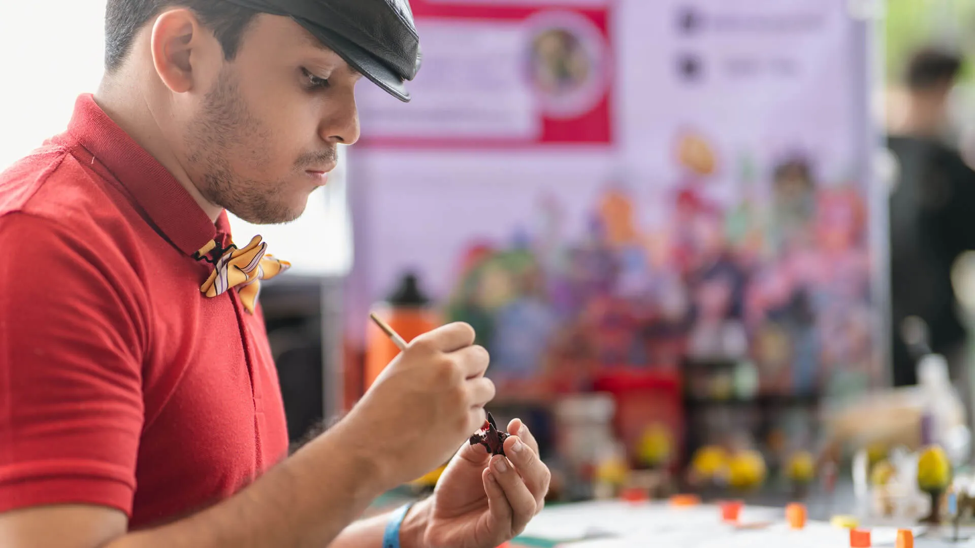 A man in a red polo shirt and newsboy cap carefully paints a small miniature figure.