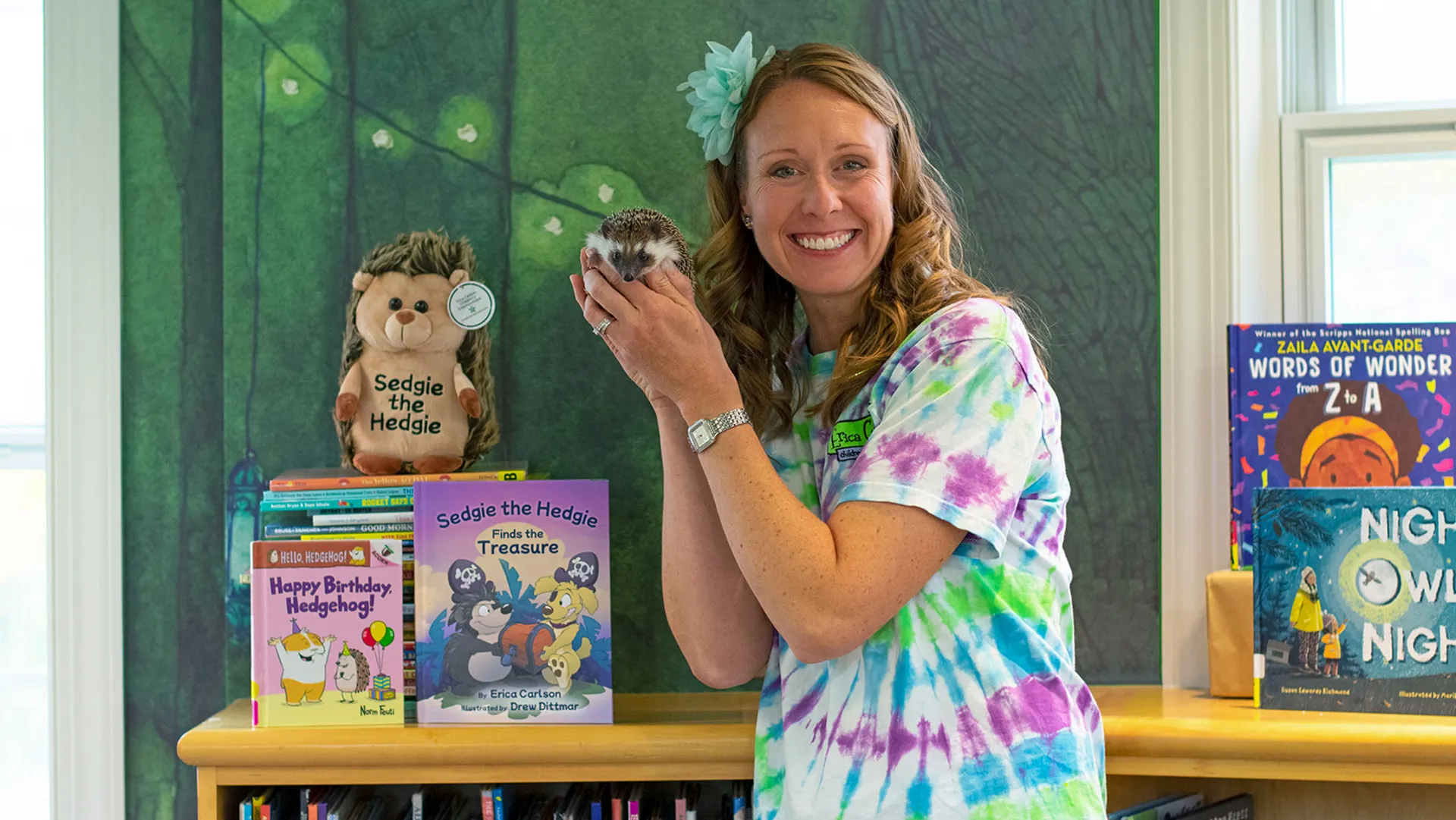 A woman in a tie-dye shirt holds a live hedgehog in front of books featuring a hedgehog character.