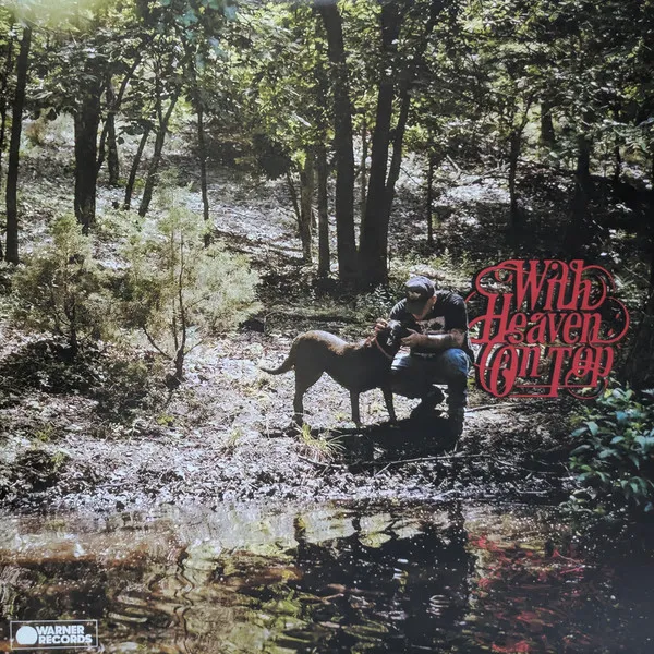 A man kneeling and petting a dog in a forest on a muddy river bank. Album cover for Zach Bryan: "With Heaven On Top."