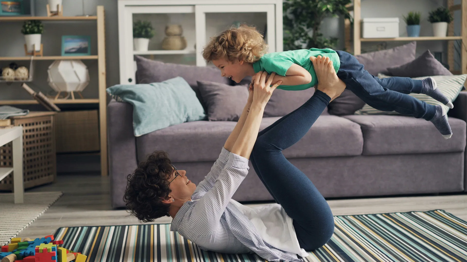 A mother lying on the floor lifts her son into the air with her feet in a living room.